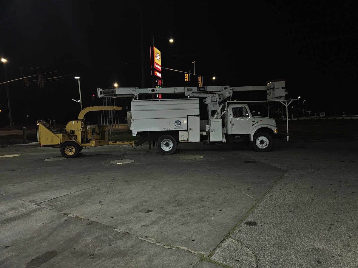A white bucket truck with a chip box and a yellow wood chipper parked on a lit lot at night.